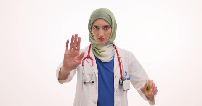 Muslim woman doctor shows caution gesture raising CBD oil. Female physician in hijab makes Stop sign with hands showing marijuana oil as narcotic