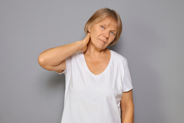 Worried mature woman wearing white T-shirt feeling pain in her neck and back showing signs of physical stress and anxiety dealing with her acute injury standing against gray background