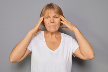 Frustrated mature woman wearing white T-shirt grimacing in pain holding head due to headache and...