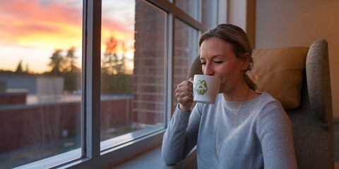 Fototapeta premium A modern woman enjoys her morning coffee, gazing thoughtfully out of the window. The soft morning light enhances the peaceful atmosphere, making this a calm moment before the day's rush.