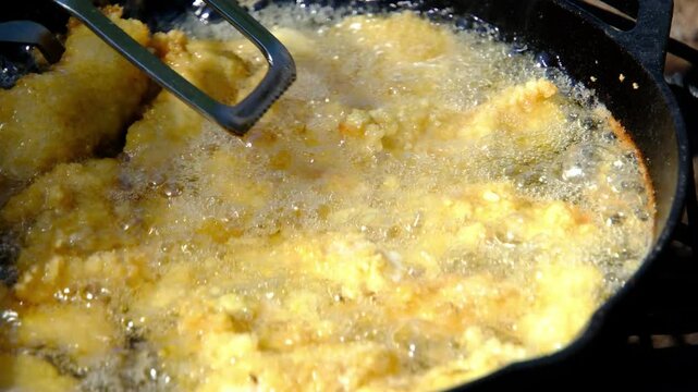 Super close up while using tongs to flip deep frying catfish nuggets or fish in batter and breadcrumbs in cast iron skillet or pan over firepit while camping outdoors