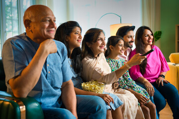 Multigenerational Indian family of six on a sofa, watching TV, sharing popcorn in joyful excitement