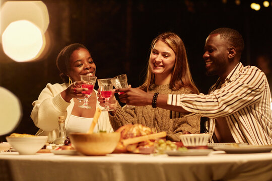 Group of three adult friends clinking glasses at table enjoying dinner outdoors in cozy low light setting with lens flare