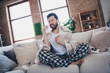 Photo of sick young man wearing sleepwear staying home feeling unwell blowing nose napkin indoors...