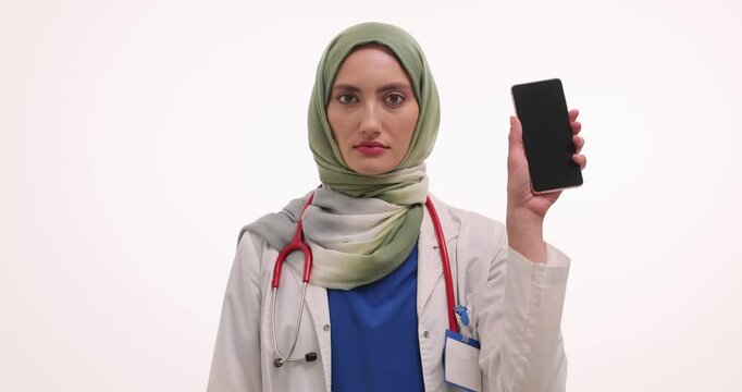 Woman doctor in hijab raises smartphone showing thumbs down. Muslim female physician makes negative nod with dislike gestures prohibiting call at procedure