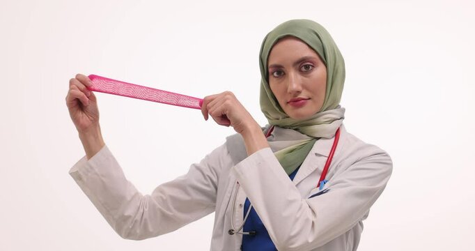 Woman doctor in hijab shows pink elastic condom on white. Muslim physician demonstrates contraceptive method for college students focusing on flexibility