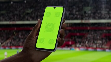GREEN SCREEN CHROMA KEY A hand holding a phone with a green screen at a stadium during a football match. Ideal mockup for sports, betting, or event apps