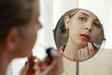 Woman gently applying red lipstick while gazing into round mirror, surrounded by serene...