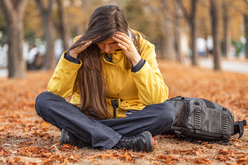 In park full of autumn leaves young woman sits cross-legged on the ground. Wearing yellow jacket. she rests her head on her hands. Concept: mental health issues stress anxiety. autumn melancholy  © Елена Бабанова