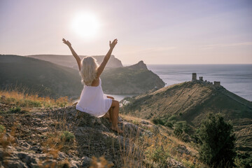 Happy woman is sitting on a hillside, wearing a white dress. She is surrounded by a beautiful landscape, with a body of sea in the background. Concept of peace and happiness.