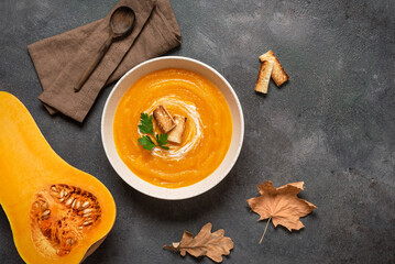 Pumpkin cream soup with croutons in a bowl and a fresh half of butternut pumpkin on dark grunge background, top view, autumn warming soup, vegetarian food