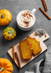 Slices of pumpkin sweet bread with cinnamon on a wooden board on a dark background with decorative pumpkins and a cup of hot chocolate or coffee.