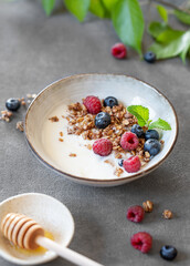 Granola with greek yogurt, raspberries, blueberries and honey in a bowl on a dark background with green leaves.