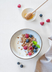Flat lay of granola with yogurt, raspberries, blueberries and honey in a bowl on a white background with natural morning light.