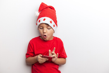 a boy in a Santa costume holds a smartphone in his hands. an emotional little santa. a cheerful child for Christmas with a phone.
