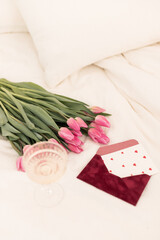 A romantic setting featuring pink tulips, a heart-decorated envelope, and a glass of sparkling drink on a soft white bedspread