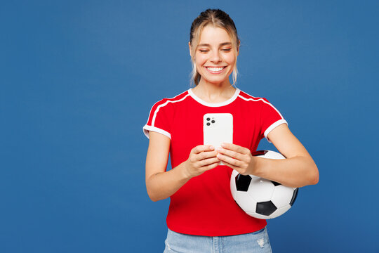 Fototapeta Young excited satisfied woman fan wearing basic red t-shirt cheer up support football sport team hold in hand soccer ball watch tv live stream use mobile cell phone isolated on plain blue background.