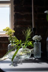 Delicate glass vases adorned with fresh flowers bathe in warm sunlight near a rustic brick wall