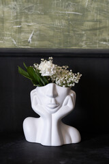 A creative ceramic planter shaped like a face, adorned with delicate white flowers, sitting against a textured green wall