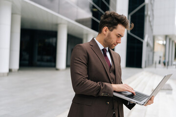 Fototapeta premium Focused businessman in suit standing outside modern office building working typing on laptop with serious expression. Handsome business man working remote online standing urban setting.