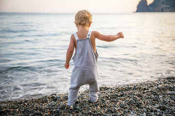 Faceless blond baby one year old in summer grey camisole stands on pebbles alone seashore in summer