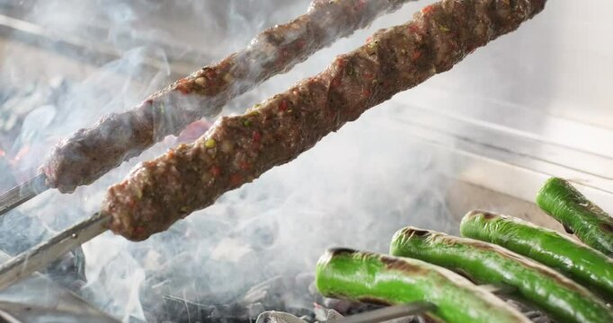 Chef prepares traditional Turkish Adana kebab, kebab grilling on skewers with vegetables tomatoes and green spicy peppers on barbecue charcoal grill