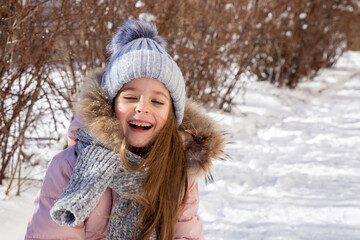 Happy laughing little girl in warm pink coat and grey knitted hat has fun and walking in snowy...