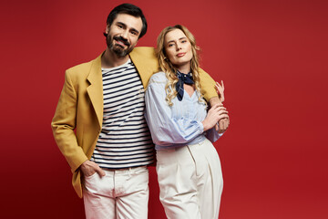 A stylish middle aged couple poses confidently in matching outfits against a bold red backdrop.