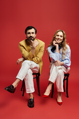 A middle aged couple showcases their fashion sense, seated gracefully on black chairs with flair.