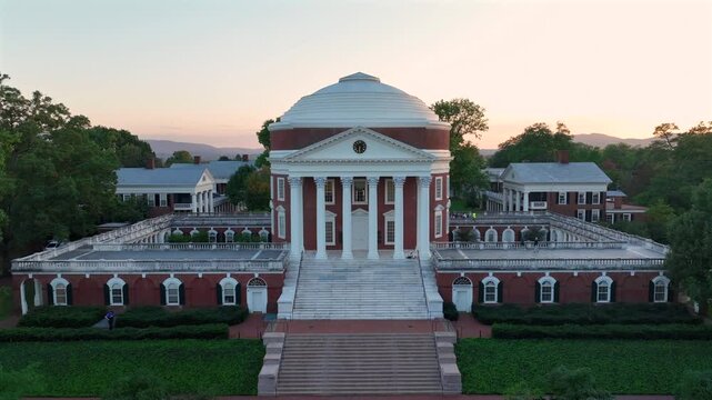 Historic The Rotunda Building with cupola at University of Virginia during golden sunset. Park area with grass and ancient faculties. Aerial rising wide shot panorama.