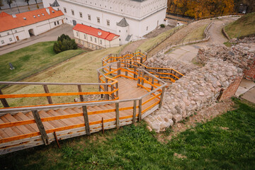 A zigzagging wooden staircase with stone walls leads down a hill toward historic buildings with red...