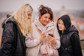 A group of three people, two girls and a woman, are smiling and looking at a phone screen together outdoors, wearing warm jackets..