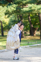 A Korean couple, a man in his 30s and a woman in her 20s, are wearing Hanbok and hugging each other in a park surrounded by green trees in Seoul, South Korea.