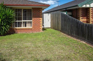 Fototapeta premium Background texture of a suburban front yard garden of a brick house in Australia. The vacant grass lawn is bordered by a wooden boundary fence, separating the property from a neighboring home.