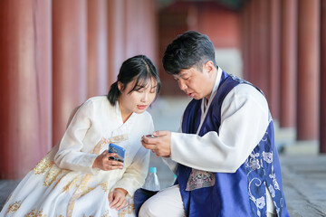 A Korean couple, a man in his 30s and a woman in her 20s, are happily wearing Hanbok and sitting in a historical building in Seoul, South Korea.