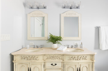 A bathroom detail with a cream cabinet and mirrors, a plant and towel sit on the marble countertop, and chrome light fixtures are mounted above the mirrors.