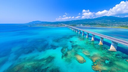 Obraz premium An aerial view of a long bridge stretching across a turquoise ocean with coral reefs visible beneath the surface. The sky is bright blue with fluffy white clouds.