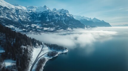 Fototapeta premium An aerial view of a winding road along the shore of a lake with snow-capped mountains in the background. The lake is shrouded in mist, creating a serene and picturesque scene.