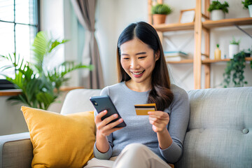 Happy asian woman buying electronic services  marketplaces e-commerce application using card and smartphone sitting sofa at home