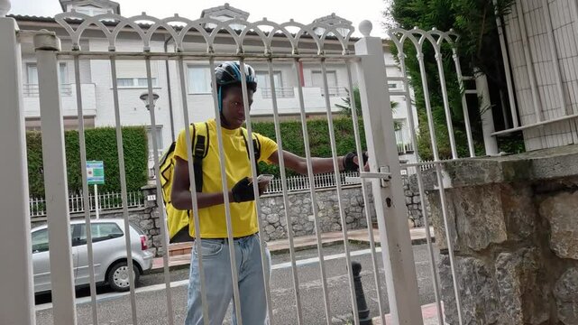 Young African delivery cyclist opening a customer gate while holding food. Concept of reliable food delivery service.
