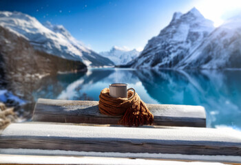 A steaming cup of coffee wrapped in a scarf sits on a wooden railing overlooking a snow-capped mountain range reflected in a calm blue lake.