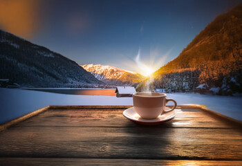 A steaming cup of coffee rests on a wooden table, overlooking a picturesque winter landscape with snow-capped mountains and a frozen lake.