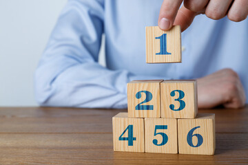 Man building pyramid of numbered cubes at wooden table, closeup. Step-by-step concept