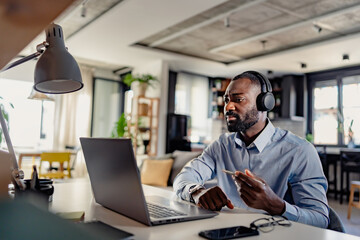 African-American businessman wearing headphones, engaged in a video call on his laptop in a modern home office. Focused and expressive during a virtual meeting.