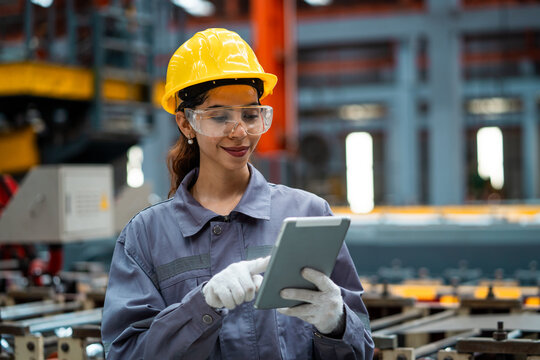 A woman wearing a yellow helmet and safety glasses is looking at a tablet. She is wearing a grey jacket and gloves