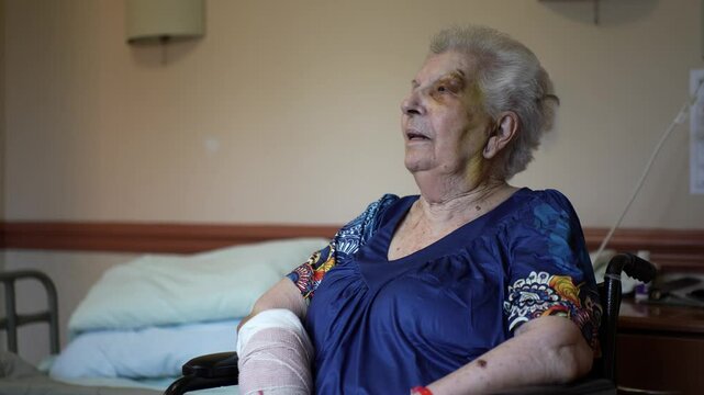 Elderly woman in muumuu and cast on arm with bruised face sitting in wheelchair in rehabilitation facility.