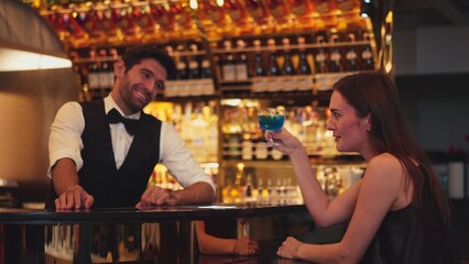 Sitting in smiling beautiful woman guest lively taking with smart bartender while drinking mixed vodka in blue hawaii cocktail and whiskey at luxurious counter bar on Friday meeting at club. Vinosity.