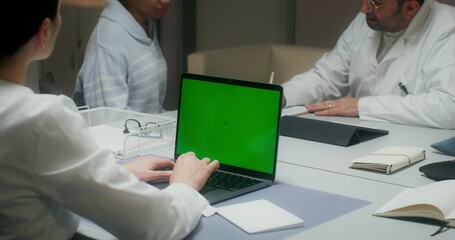 The nurse uses a laptop with a green screen, entering patient data. A woman talks to a doctor sitting at a table in the background