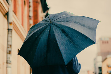 It's raining in the city and a man in a blue cardigan is walking under a blue umbrella as drops of...