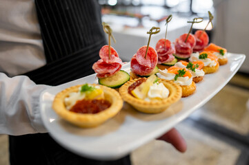 A close-up of a hand holding a tray with assorted canapés, featuring toppings like cured meats, salmon, cream cheese, and cucumber. blurred background emphasizes the vibrant, appetizing presentation